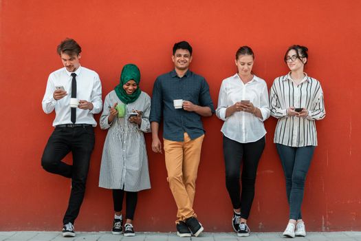 Multiethnic group of casual businesspeople using smartphones during a coffee break from work in front of the red wall outside. High-quality photo