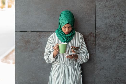 African Muslim businesswoman with green hijab using a smartphone during a coffee break from work outside. High-quality photo