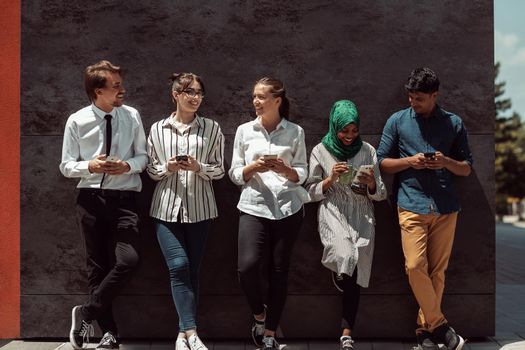 Multiethnic group of casual businesspeople using a smartphone during a coffee break from work in front of the black wall outside. High-quality photo