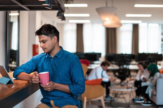 Casual businessman taking a break from the work using a laptop while drinking tea in relaxation area of modern open plan startup office. High-quality photo