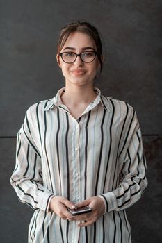 Startup businesswoman in shirt with a glasses using smartphone while standing in front of gray wall during a break from work outside. High-quality photo