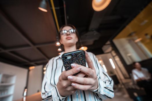 Businesswoman with glasses using a smartphone at modern startup open plan office interior. Selective focus. High-quality photo