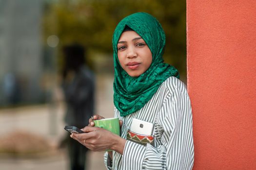 African Muslim businesswoman with green hijab using a smartphone during a coffee break from work outside. High-quality photo