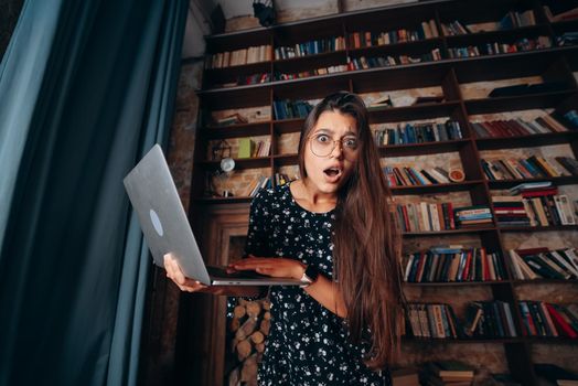 Young beautiful woman in eyeglasses looks shocked after checking mail on laptop and looking at the camera