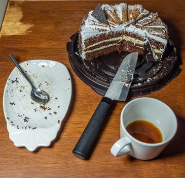 White plate with crumbs and a spoon after the cake, drinking a cup of coffee and a cut cake on a wooden table, top view.