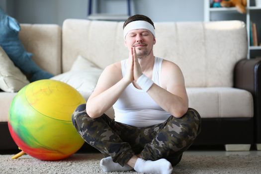 Man with praying hands and closed eyes meditates in lotus position at home. Exercises for relaxation and relieving emotional and muscle tension