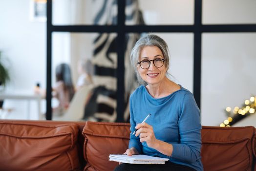 Happy senior woman in glasses writing to notebook or diary at home