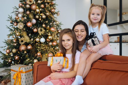 Happy mother and two little daughters are sitting on sofa and smiling against background of Christmas tree