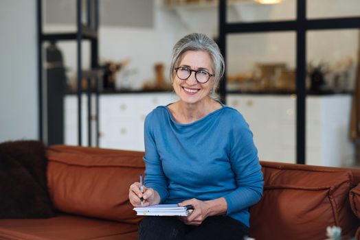 Happy senior woman in glasses writing to notebook or diary at home