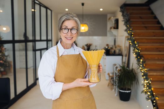 Happy senior woman showing pasta on camera. Portrait of smiling aged lady holding spaghetti standing in modern kitchen