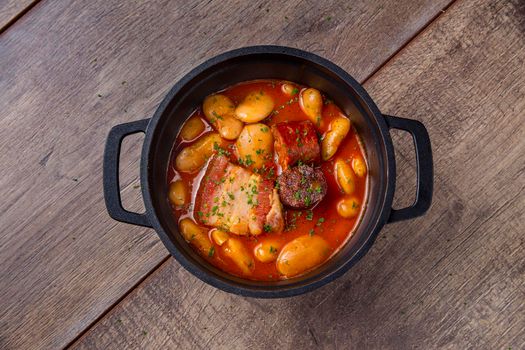 Overhead shot of a still life of white beans stewed with bacon and chorizo in an iron casserole on a wooden board. Concept of traditional Spanish gastronomy.