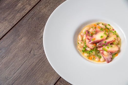 Still life of octopus with white beans and vegetables on a white plate on a wooden board. Concept of traditional Spanish gastronomy.