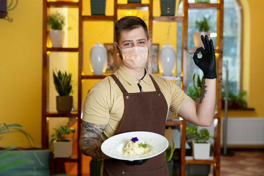 Male waiter in a protective medical mask holds a plate of pasta in his hands.