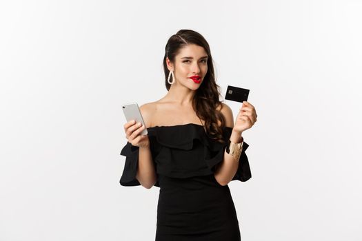 Fashion and shopping concept. Attractive woman with red lips, black dress, thinking what to buy, holding credit card and mobile phone, standing over white background.