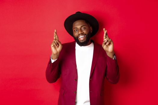 Hopeful african american man making wish, holding fingers crossed for good luck and smiling optimistic, standing against red party background.