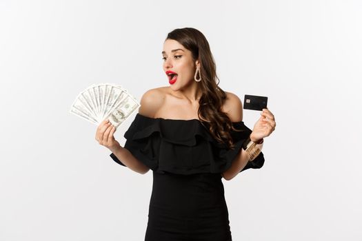 Beauty and shopping concept. Fashionable woman holding credit card, looking at money with amazement, standing over white background.