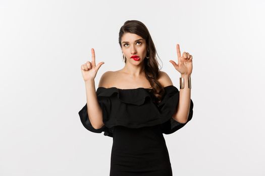 Fashion and beauty. Thoughtful attractive woman in black dress staring and pointing up, thinking with tempted look, white background.