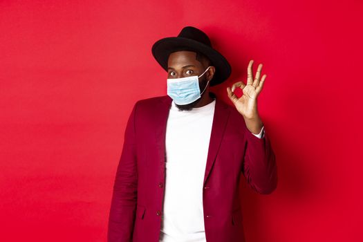 Covid-19 and fashion concept. Stylish african american man in hat and blazer, wearing face mask and showing okay sign, standing over red background.