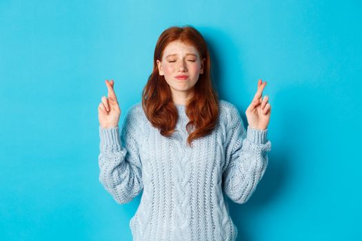 Hopeful redhead girl making a wish, cross fingers for good luck, smiling and anticipating good news or positive result, standing against blue background.