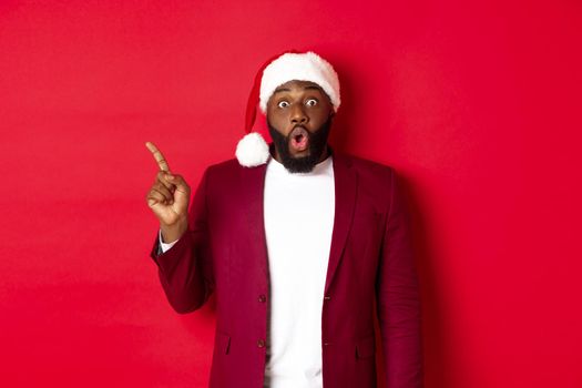 Christmas, party and holidays concept. Impressed Black man with beard, wearing santa hat, pointing finger left and gasping amazed, standing against red background.