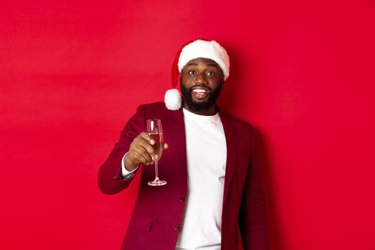 Christmas, party and holidays concept. Cheerful Black man saying cheers, raising glass of champagne and wishing happy New Year, standing against red background.