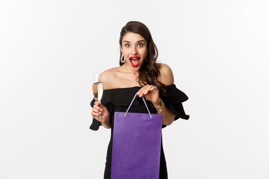Excited woman receiving christmas gift in shopping bag, holding glass of champagne and looking surprised at camera, standing over white background.