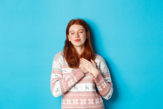 Image of dreamy redhead girl close eyes and holding hands on heart, feeling nostalgic, remember or imagine something, standing over blue background.