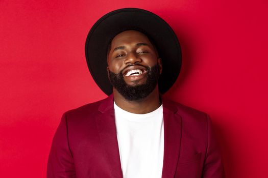 Close-up of happy handsome Black man smiling at you, looking pleased, wearing black hat and blazer, red background.