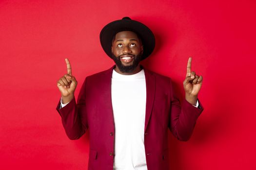 Winter holidays and shopping concept. Happy african american man wearing party clothing for new year, pointing and looking up with pleased smile, red background.