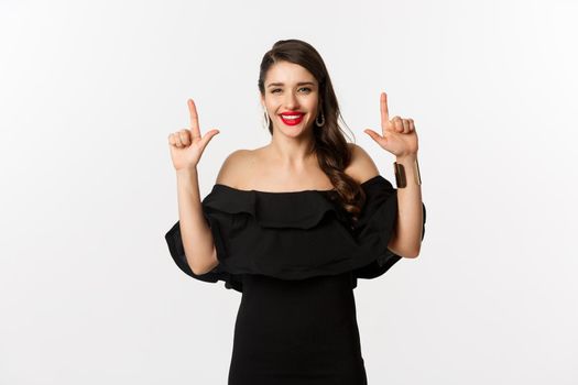 Fashion and beauty. Charming woman with red lips, black dress, smiling happy and pointing fingers up, showing logo, white background.