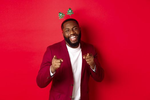 Merry Christmas. Handsome african american man in blazer and party headband, celebrating new year, pointing at camera and smiling, congratulating, red background.