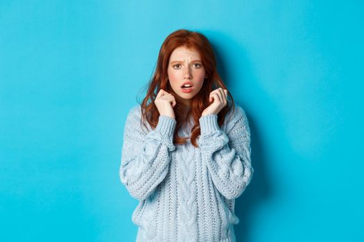 Image of scared teenage girl with red hair, jumping startled and looking alarmed, standing over blue background.