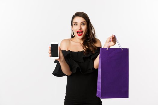 Beauty and shopping concept. Beautiful and stylish woman showing smartphone screen and bag, buying online, standing over white background.