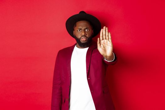 Serious and concerned Black man stretching hand to stop you, prohibit and disagree, frowning and grimacing displeased, standing over red background.