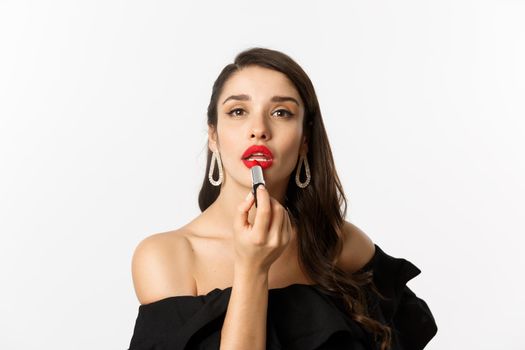 Close-up of beautiful woman applying red lipstick on lips, looking at camera like mirror, standing in black dress over white background.