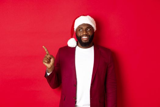Christmas, party and holidays concept. Cheerful Black man smiling, pointing fingers left and looking at logo, standing over red background.