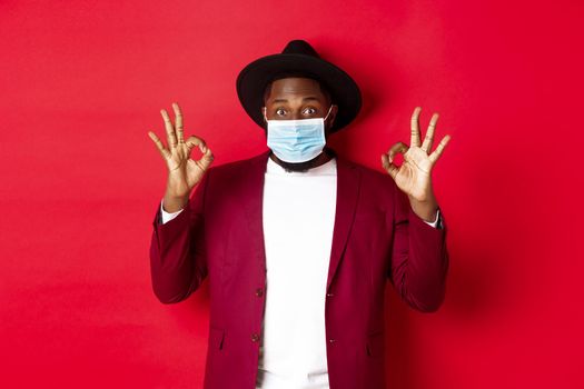 Covid-19 and fashion concept. Stylish african american man in hat and blazer, wearing face mask and showing okay sign, standing over red background.