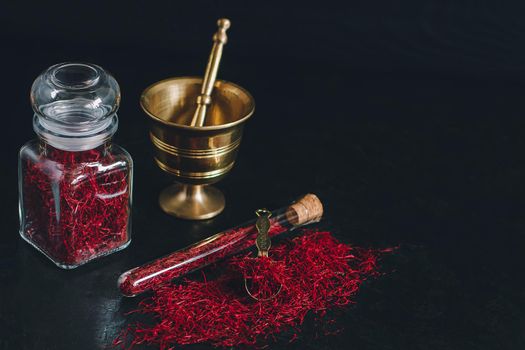 Raw organic red dried saffron spice on wooden background in vintage metal brass mortar with pestle, glass jar and tube.