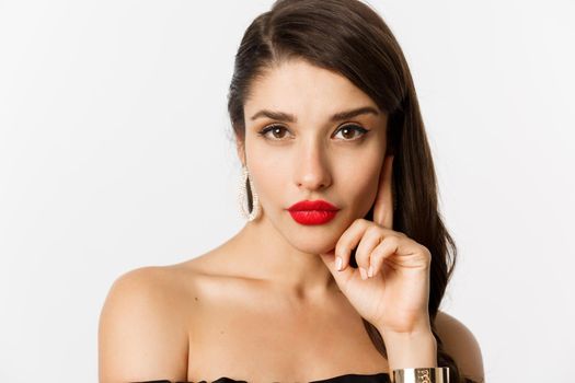 Fashion and beauty concept. Close-up of elegant beautiful woman in black dress, evening makeup and red lipstick, looking sassy at camera, standing over white background.