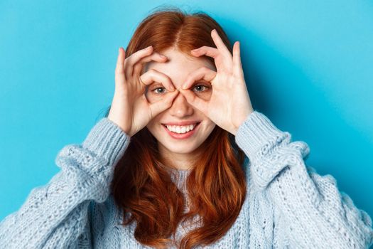 Close-up of funny and cute redhead girl making hand glasses and looking through them, seeing promo offer and smiling, standing over blue background.