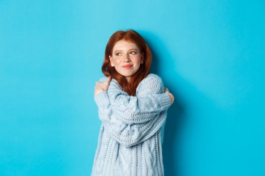 Happy cute redhead girl hugging herself, wearing comfortable and warm sweater, smiling at camera, standing over blue background.