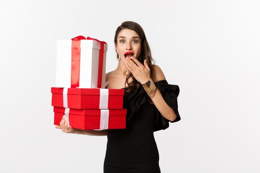 Celebration and christmas holidays concept. Woman holding xmas gifts and looking surprised, receive presents, standing over white background.