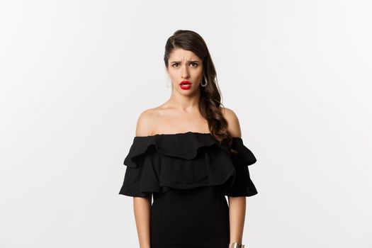 Fashion and beauty. Disappointed and upset woman in black dress staring at camera displeased, complaining with jealous look, standing over white background.