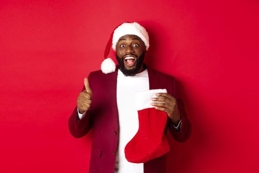 Excited Black man showing thumb-up in approval, holding christmas sock with holiday gifts, smiling amazed, standing over red background.
