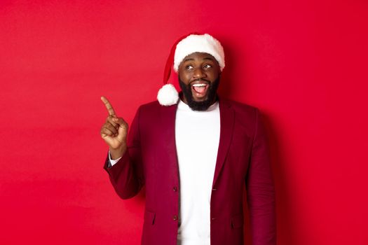 Christmas, party and holidays concept. Cheerful Black guy in santa hat pointing fingers left, showing banner, standing amused against red background.