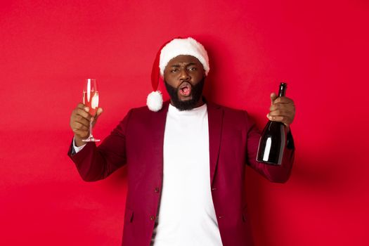 Christmas, party and holidays concept. Cheerful man enjoying New Year, wearing santa hat, raising glass and bottle of champagne, having fun against red background.
