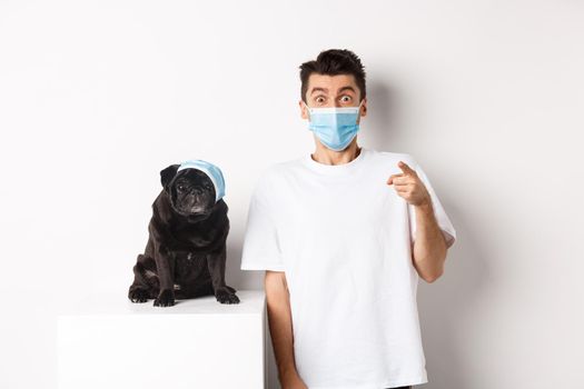 Covid-19, animals and quarantine concept. Happy dog owner and cute pug wearing medical masks, man pointing finger at camera amazed, white background.