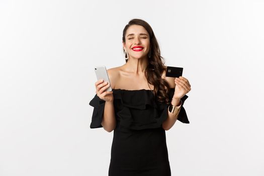 Online shopping concept. Fashionable woman in black dress, holding credit card with smartphone, looking satisfied, standing over white background.