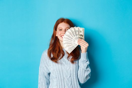 Delighted redhead woman showing money near face, smiling satisfied, standing ready for shopping over blue background.