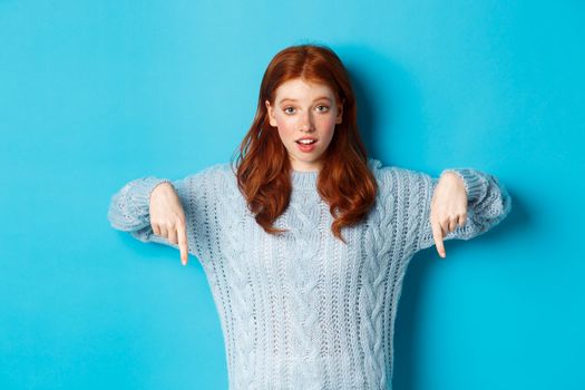 Winter holidays and people concept. Cute teenage girl pointing fingers down, showing advertisement, standing over blue background.